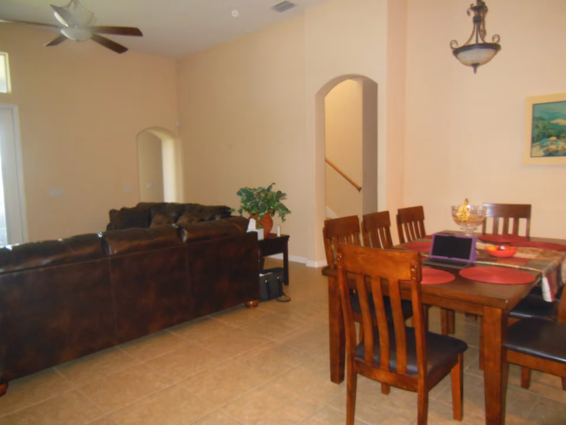 Interior view of a living and dining area with a brown leather sectional sofa, a wooden dining table with six chairs, a ceiling fan, a small side table with a plant, and a staircase visible through an arched doorway.