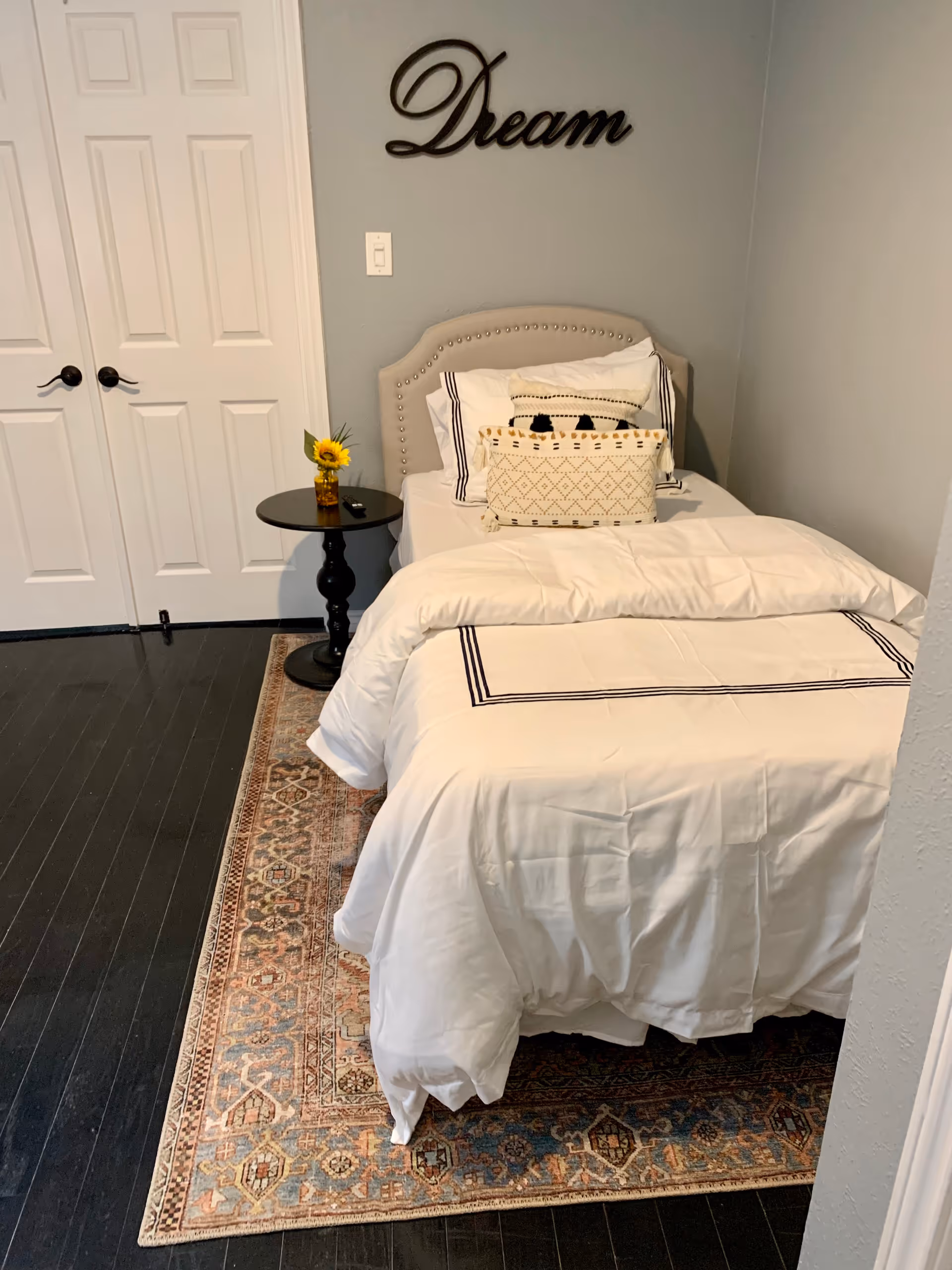 A neatly made single bed with white bedding and decorative pillows in a small bedroom. Above the bed, the word 'Dream' is mounted on the light gray wall. Next to the bed is a small round black side table with a small vase holding a sunflower. The floor is dark wood with a patterned area rug under the bed. White double doors are visible in the background.
