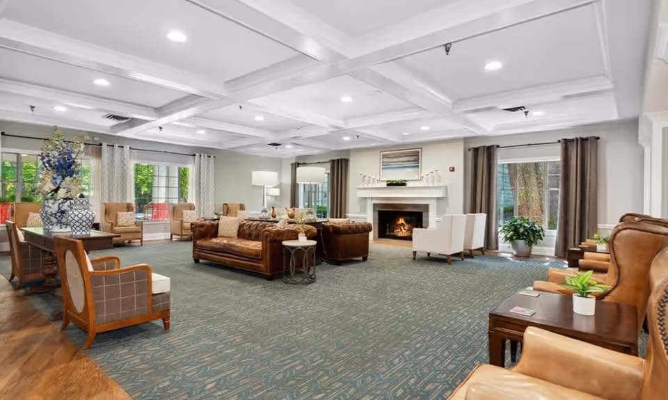 Spacious and well-lit living room area in MerryWood on Park featuring multiple seating options including leather sofas, armchairs, and wooden chairs. The room has large windows with curtains, a fireplace with a painting above it, and decorative plants and vases. The ceiling has recessed lighting and a coffered design.