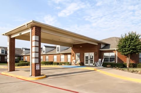 Front entrance of a single-story brick senior living facility with a covered porte-cochère and rocking chairs on the porch.
