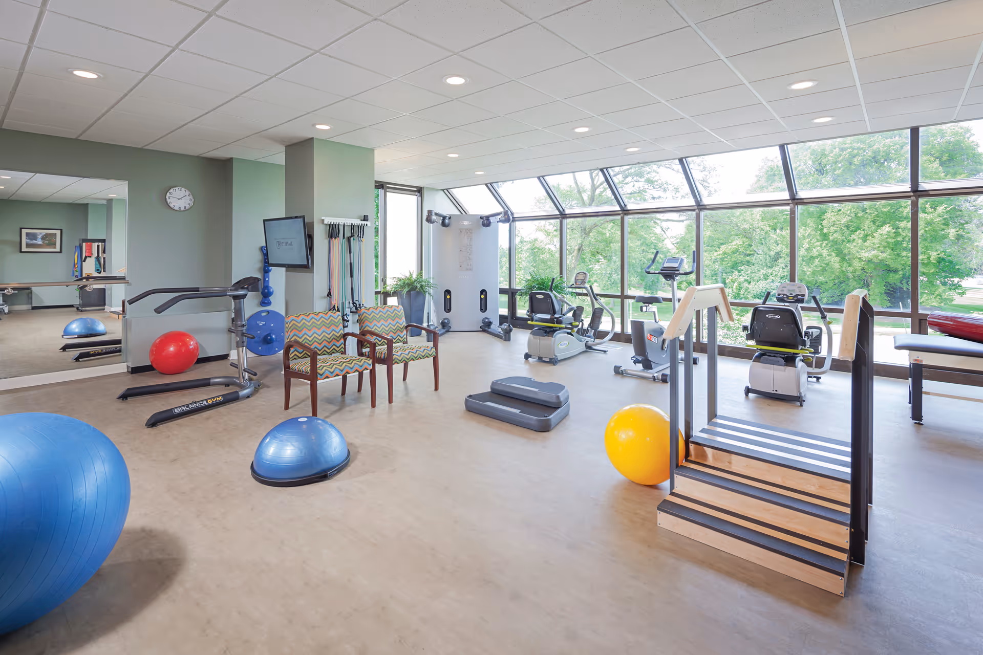 A bright and spacious fitness room with large windows overlooking greenery. The room contains various exercise equipment including stationary bikes, balance balls, a treadmill, resistance bands, and a small set of stairs for physical therapy. Two patterned chairs are placed near a wall-mounted TV and a clock. The room has a light-colored floor and a white ceiling with recessed lighting.