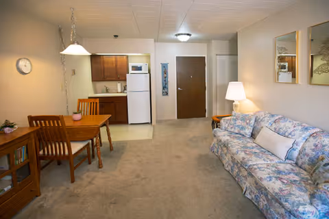 Open living area with a floral couch, wooden dining table, and a small kitchenette near the entrance door.