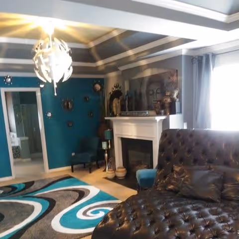 A cozy living room with a tufted dark brown leather sectional sofa, a colorful patterned rug in shades of teal, black, white, and gray, a white fireplace with decorative items on the mantel, teal accent wall with circular wall decorations, a teal armchair, and a modern white ceiling light fixture. Natural light enters through a window with gray curtains.