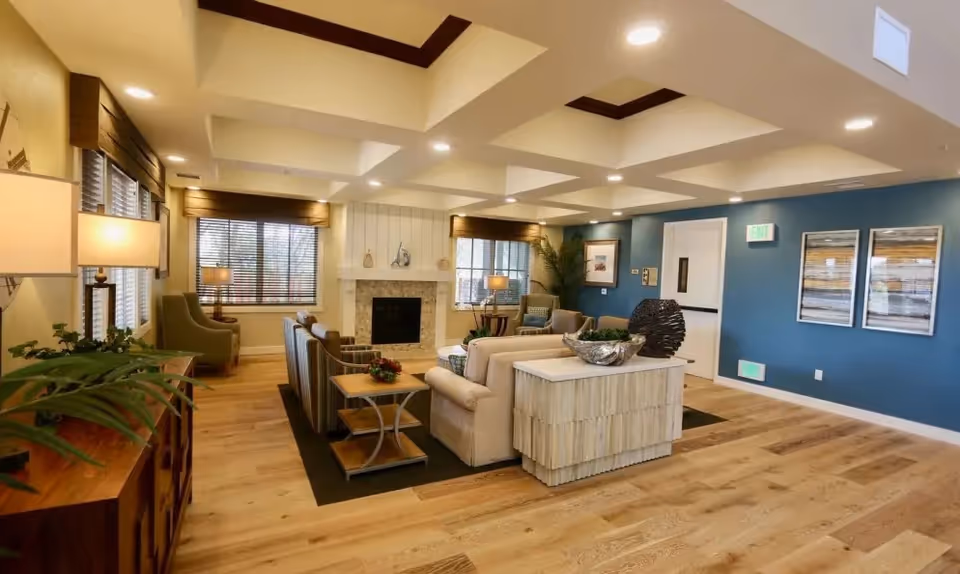 A spacious and well-lit living room area in a senior living facility with wooden flooring, beige and patterned sofas, armchairs, a fireplace, large windows with blinds, decorative plants, and modern ceiling lights.
