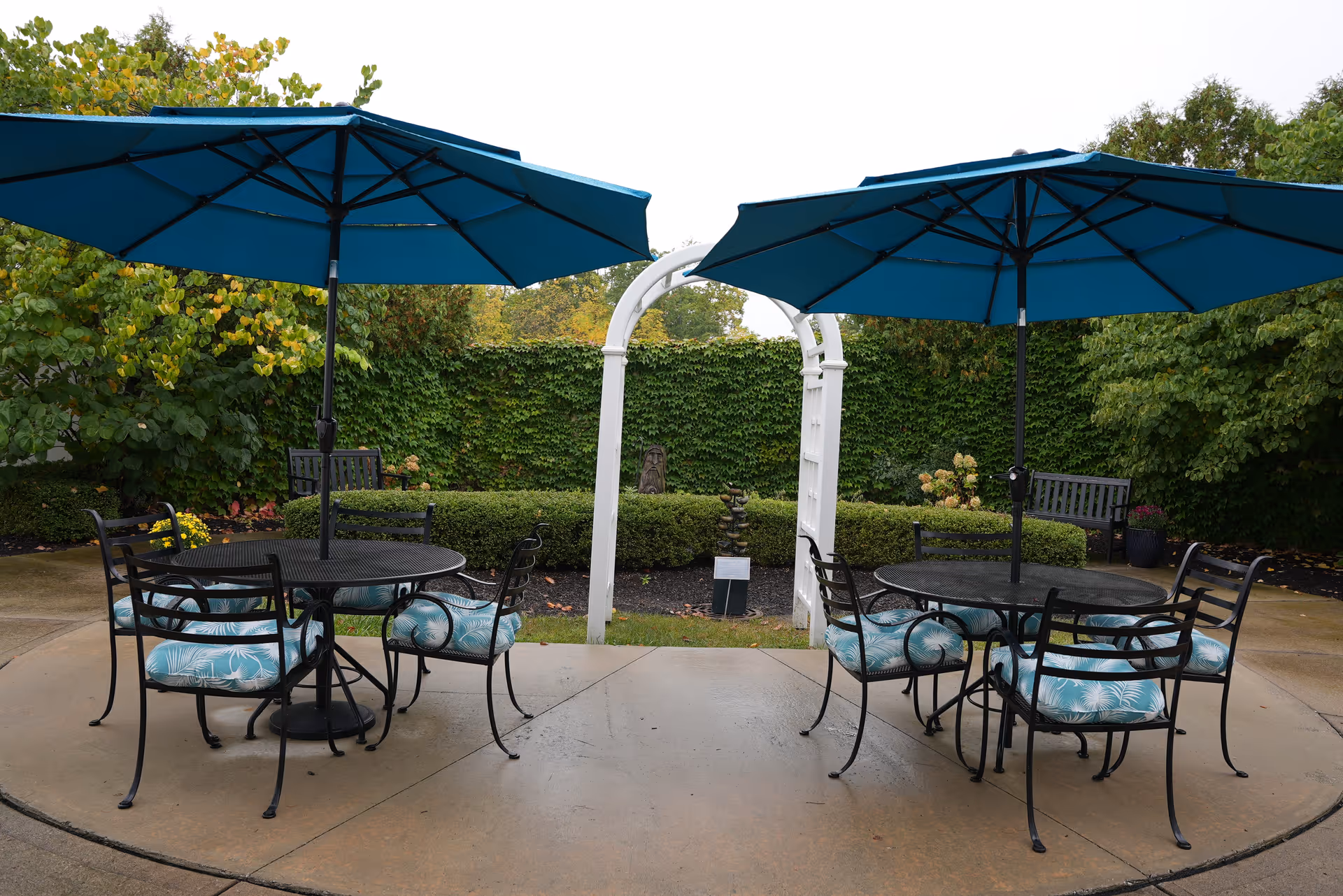 Outdoor patio with two round tables, teal umbrellas and cushioned chairs in front of a white arbor and hedged garden.