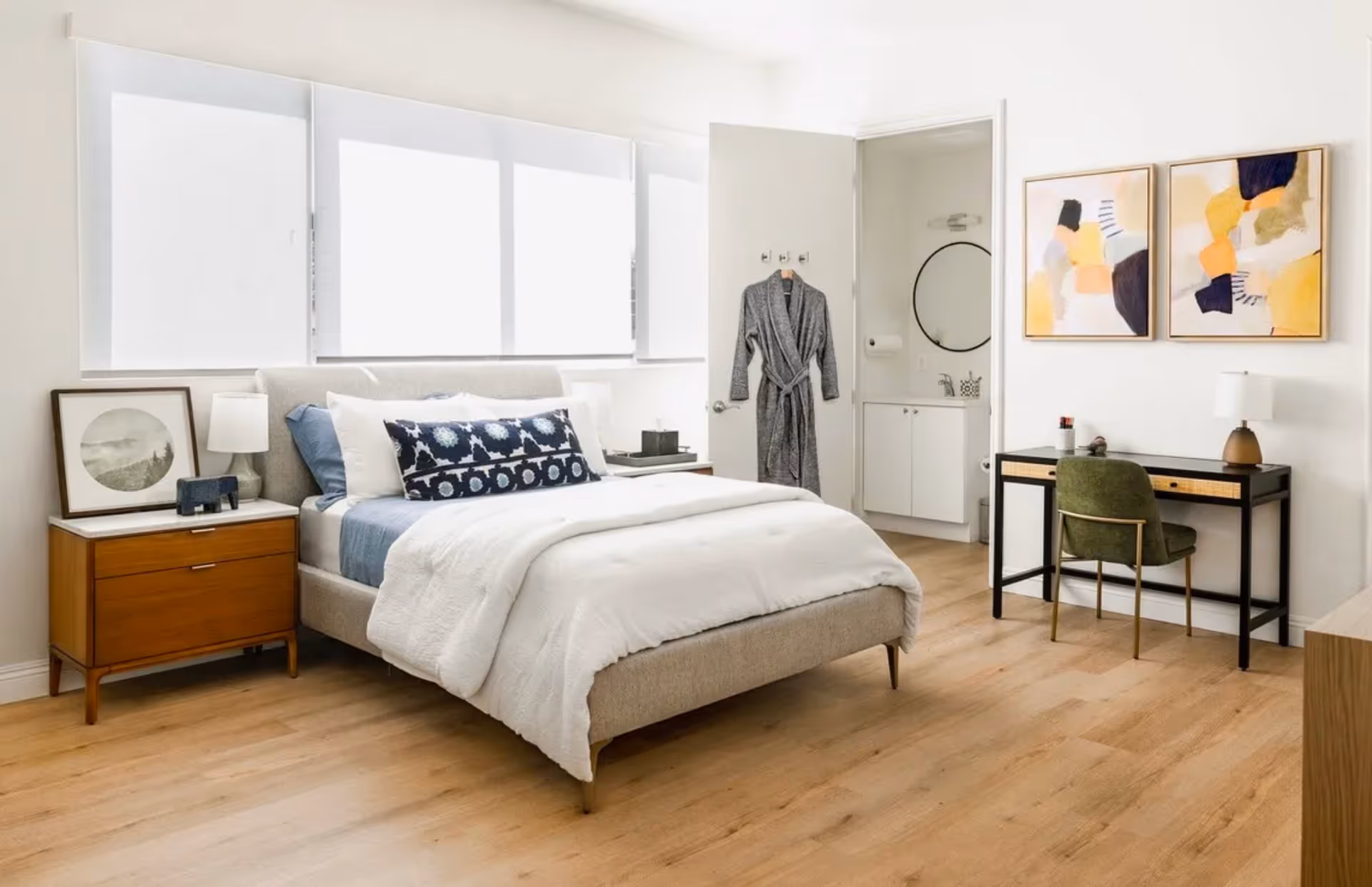 A bright and modern bedroom with a neatly made bed featuring white and blue bedding. To the left of the bed is a wooden nightstand with a lamp and framed artwork. A gray robe hangs on the open door leading to a small bathroom with a round mirror and white cabinetry. On the right side of the room is a black desk with a green chair, a lamp, and two abstract paintings on the wall above it. The room has light wood flooring and large windows with white blinds.