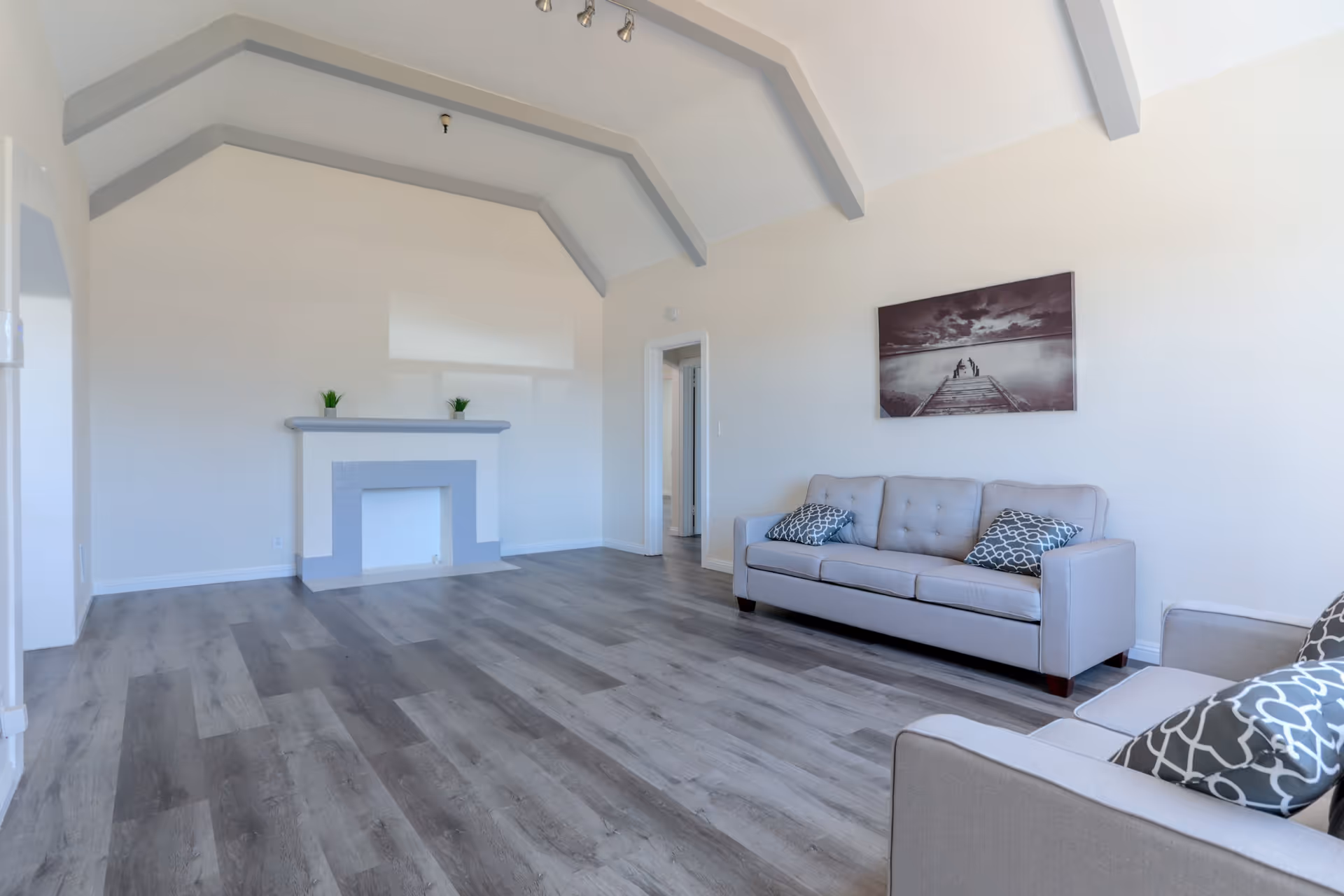 A spacious living room with light gray walls and a vaulted ceiling with exposed beams painted gray. The room features a gray sofa and an armchair with patterned cushions, a simple fireplace with a gray mantel, and a black and white photograph of a pier over water hanging on the wall. The floor is covered with gray wood-like vinyl planks.