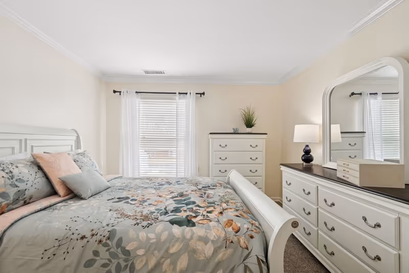A bright bedroom with a large bed featuring floral bedding and multiple pillows. There is a window with white curtains behind the bed. To the right, there is a white dresser with a large mirror, a table lamp, and a decorative box. Another white chest of drawers with a small plant on top is positioned against the far wall.