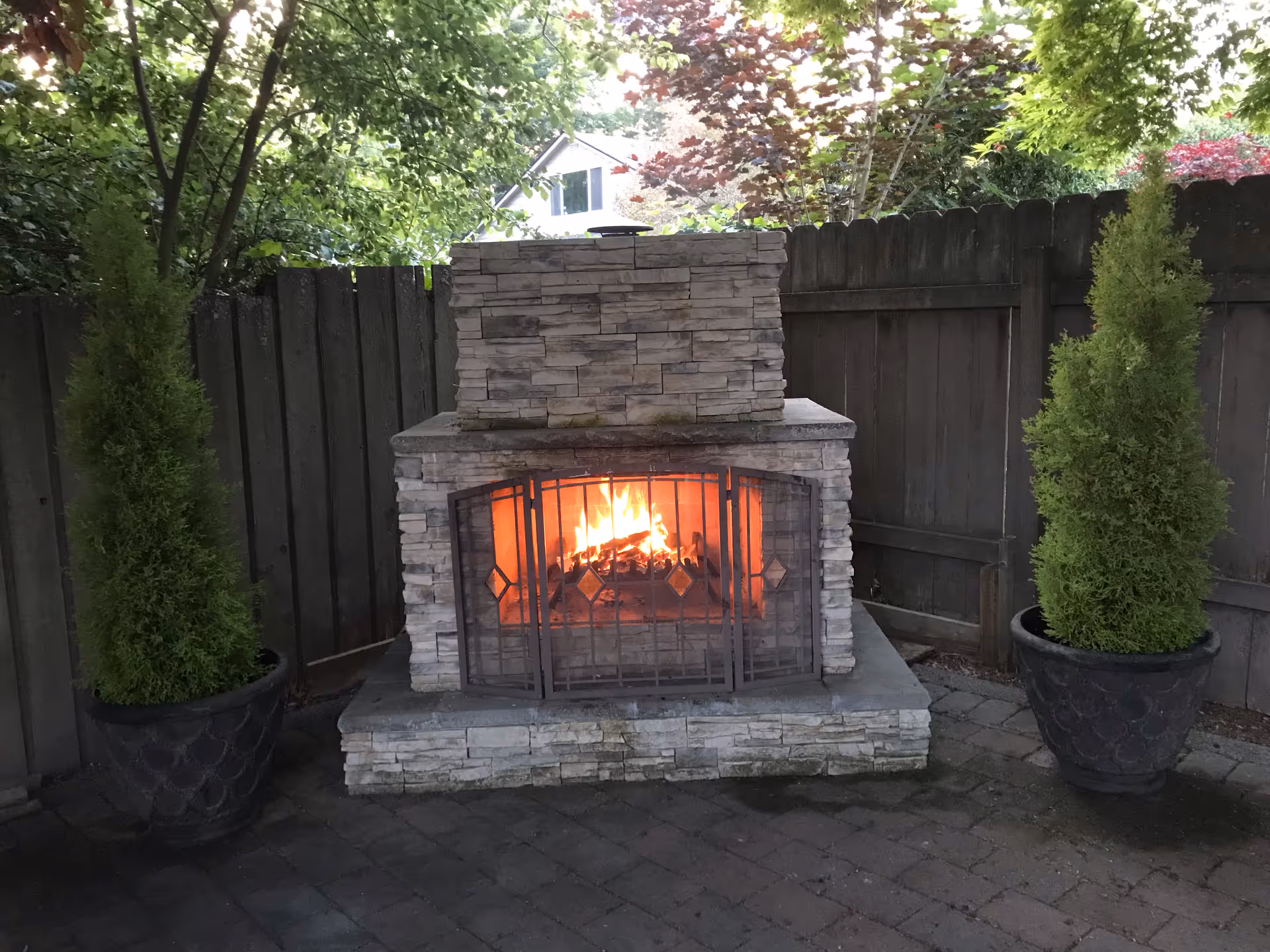 Outdoor stone fireplace with a fire burning inside, flanked by two potted evergreen shrubs, set against a wooden fence with trees and a house visible in the background.