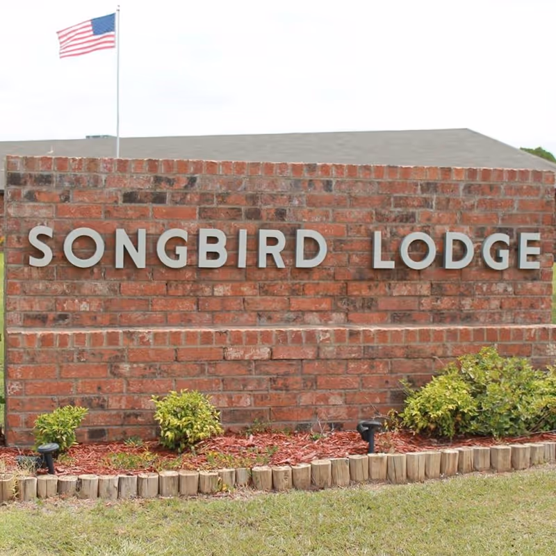 A brick sign with the words 'SONGBIRD LODGE' mounted on it, with small bushes and mulch at the base. An American flag on a flagpole is visible in the background.