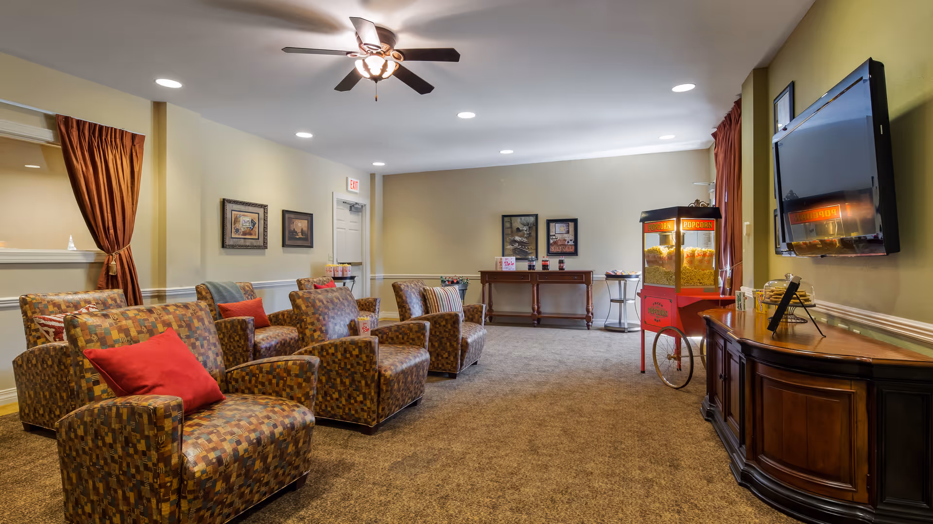 A cozy living room area with patterned armchairs each having a red pillow, a ceiling fan with lights, framed artwork on the walls, a popcorn machine, a flat-screen TV mounted on the wall, and a wooden sideboard with snacks and a photo frame.