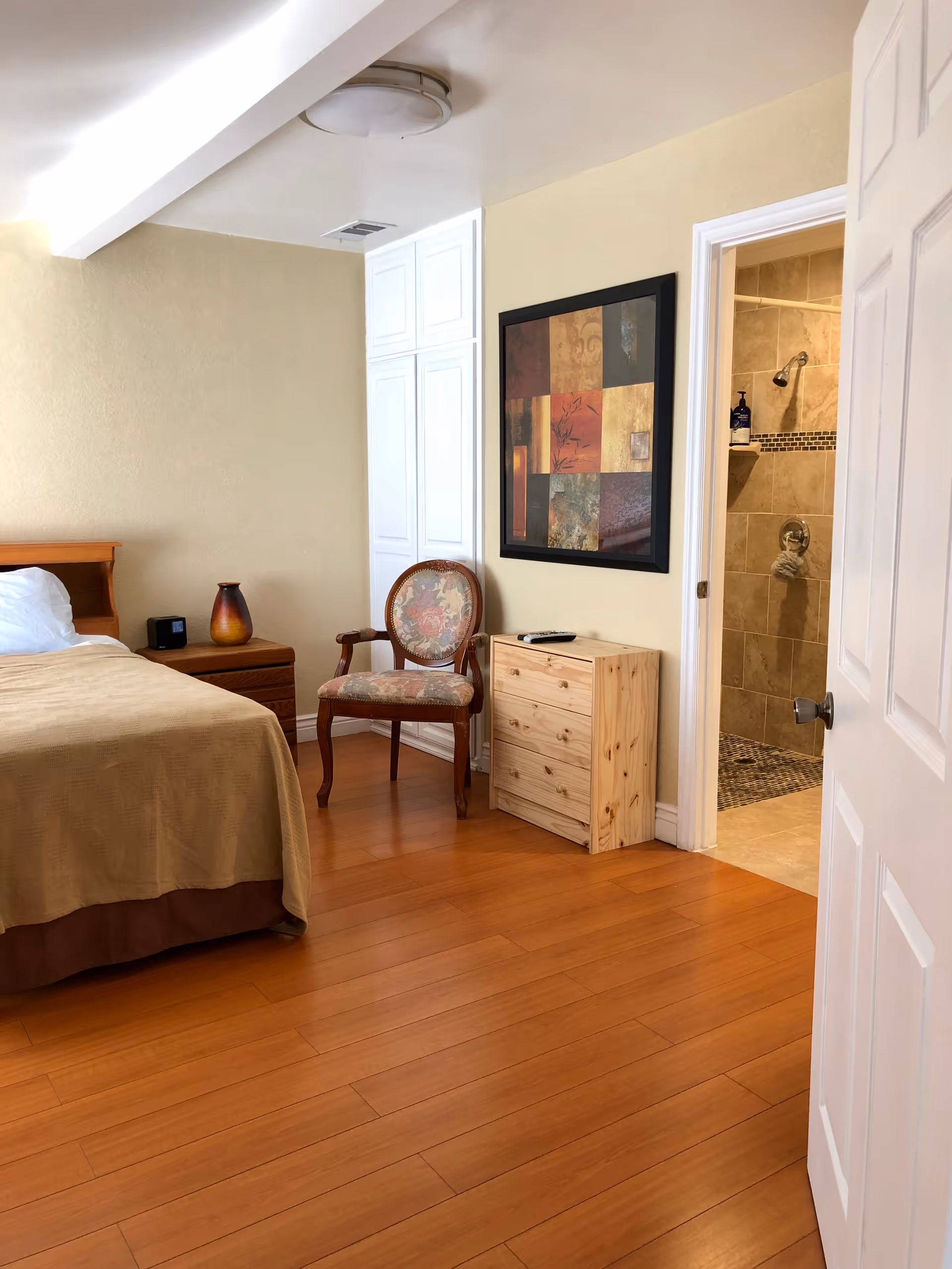 A bedroom with wooden flooring, a bed with beige bedding, a wooden nightstand with a vase and a clock, an upholstered wooden chair, a small wooden dresser, and a framed abstract painting on the wall. An open door reveals a tiled bathroom with a shower.
