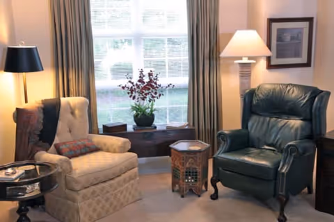 A cozy living room area with a beige upholstered armchair and a dark green leather armchair. Between them is a small decorative wooden table and a larger chest under a window with beige curtains. There are two lamps, one floor lamp with a black shade next to the beige chair and one table lamp with a beige shade behind the green chair. A framed picture hangs on the wall.