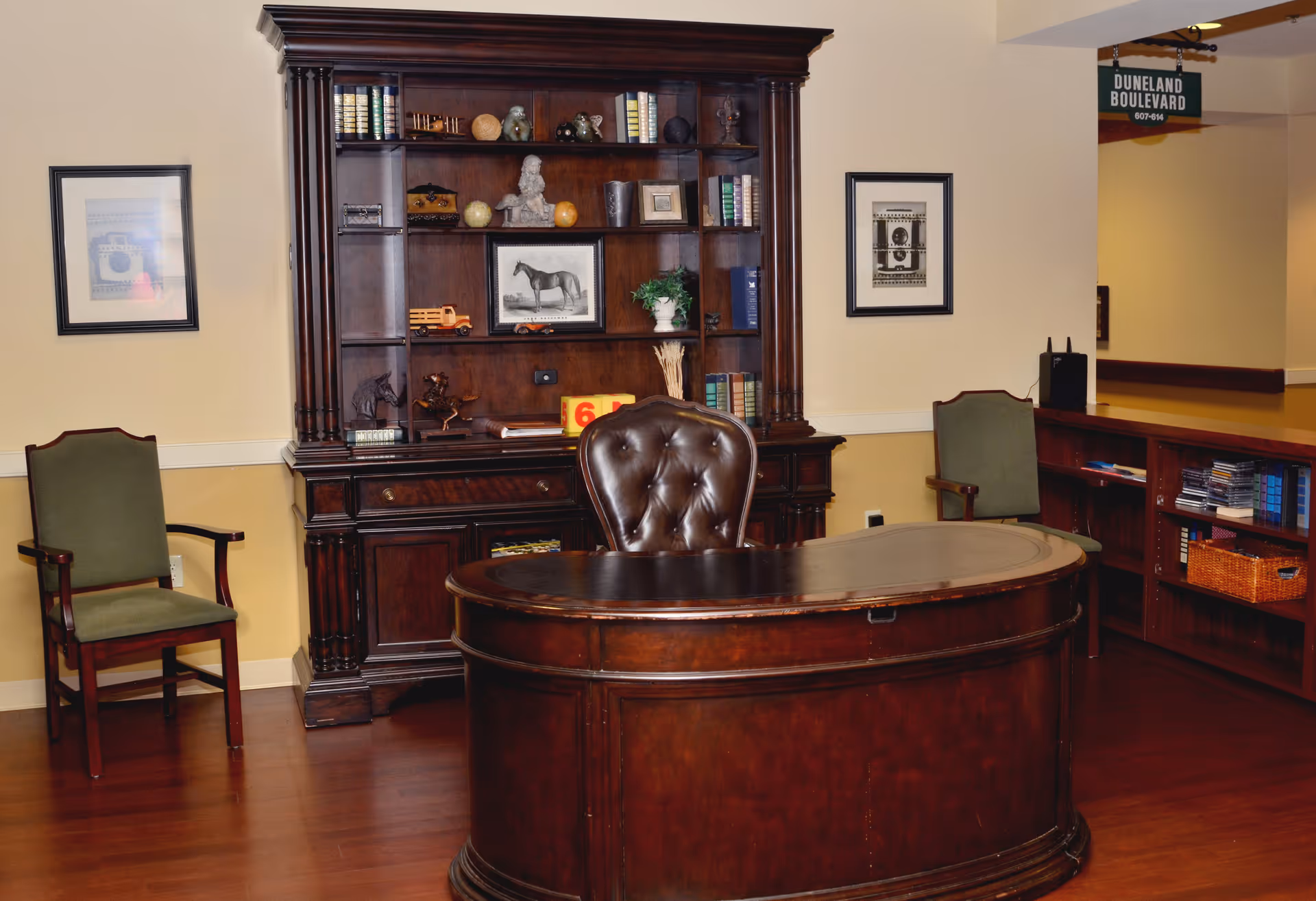 An elegant office area with a large dark wooden desk and a matching bookshelf filled with books and decorative items. Two green upholstered chairs are positioned on either side of the desk. The walls are light-colored with framed artwork, and a sign reading 'Duneland Boulevard' hangs in the background.