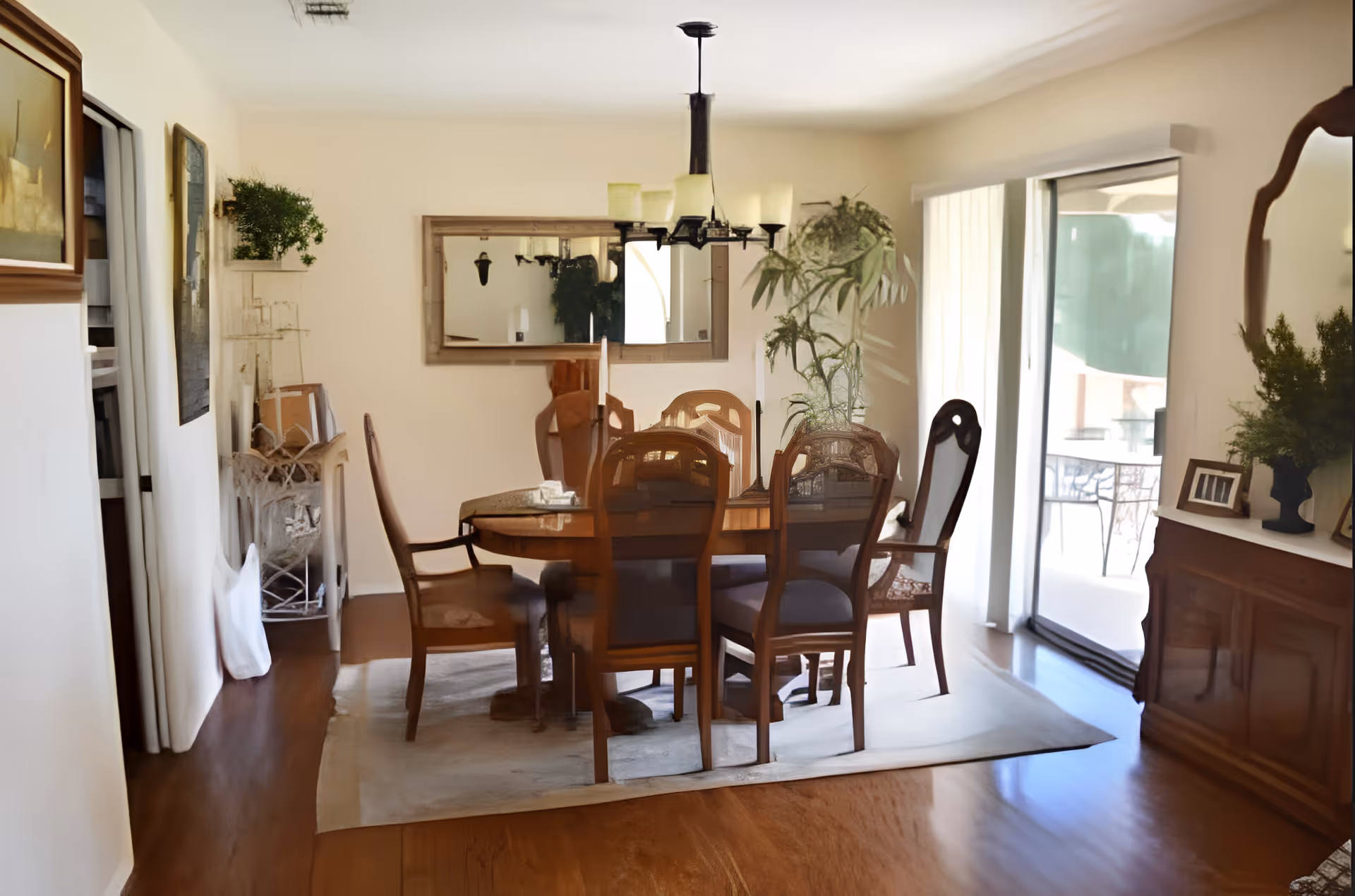 A dining room with a wooden dining table surrounded by six chairs, a large mirror on the wall, a chandelier hanging above the table, and sliding glass doors leading to an outdoor patio. The room has wooden flooring, a rug under the table, and various plants and framed pictures as decor.