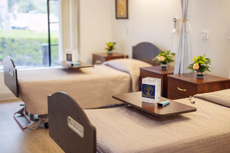 A bright room with two hospital-style beds covered with beige bedspreads. Each bed has a wooden headboard and a bedside table with a small flower arrangement. One bed has an overbed table with a tissue box and a remote control on it. A large window with a view of greenery is visible in the background.