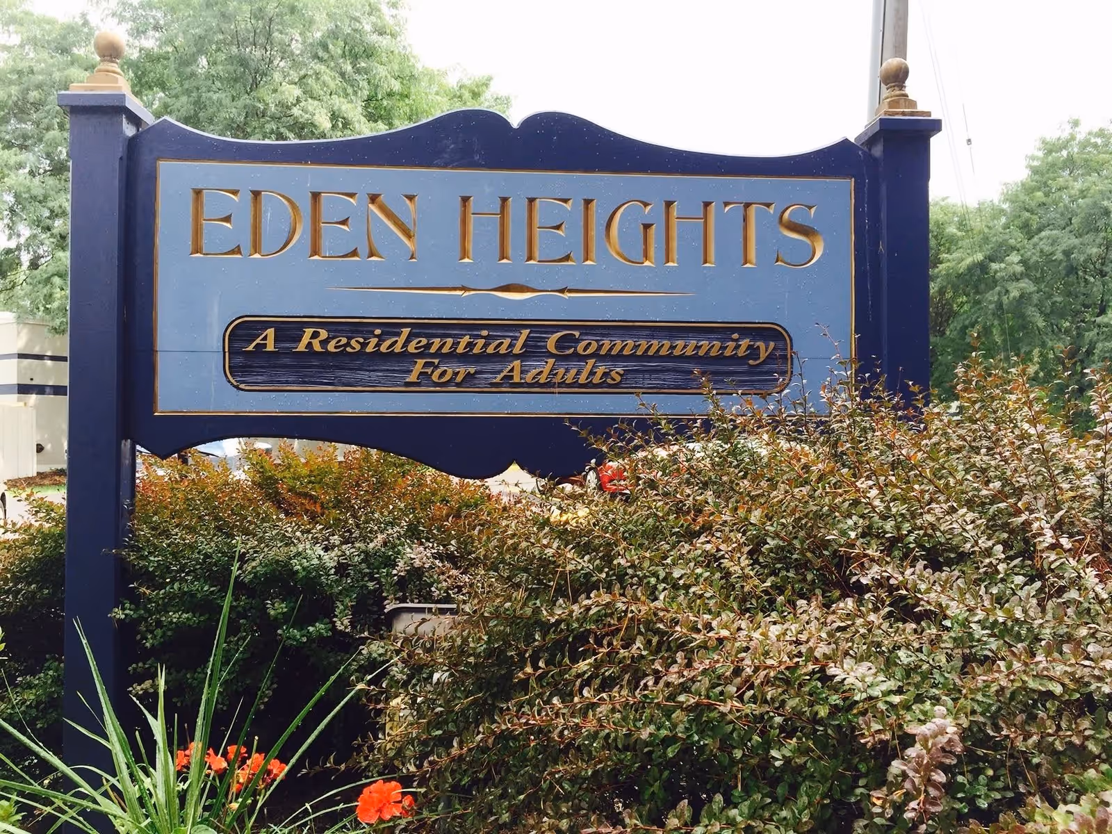 A blue and gold sign for Eden Heights, a residential community for adults, surrounded by green bushes and some red flowers with trees in the background.