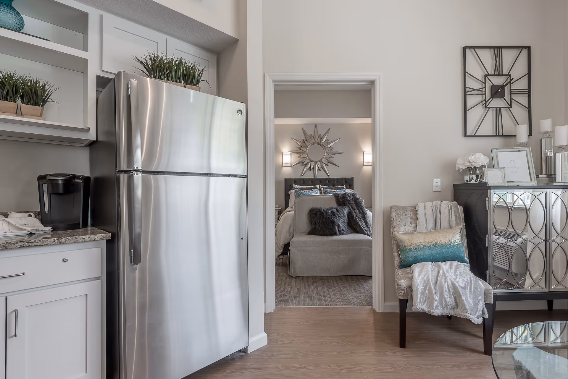 View of a modern kitchen area with a stainless steel refrigerator and a coffee maker on the counter. Through an open doorway, a bedroom is visible with a bed, decorative pillows, a sunburst mirror above the headboard, and wall sconces. To the right, there is a decorative chair with a throw blanket and pillow, a mirrored cabinet, and a large wall clock.