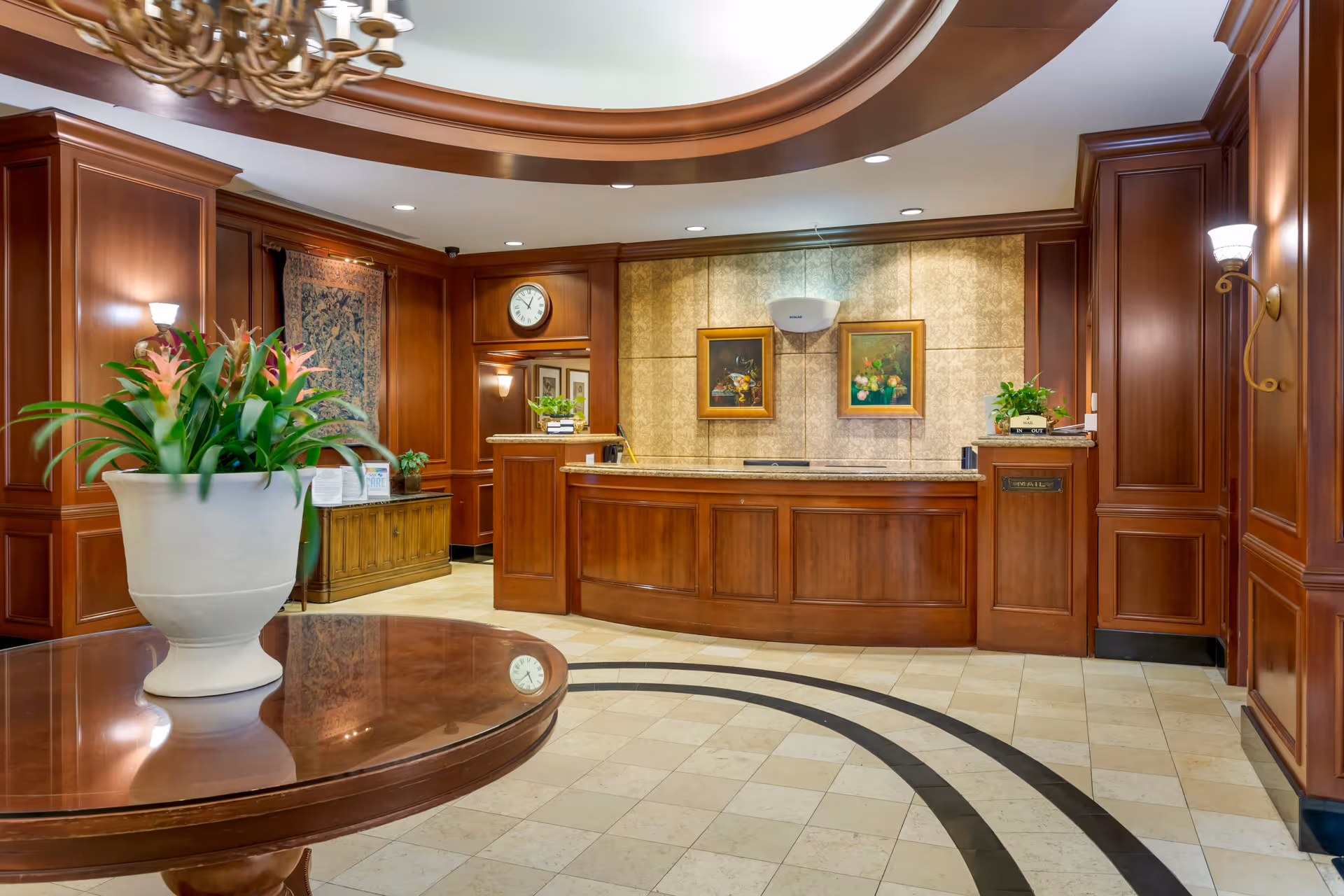 A reception area with a wooden front desk, decorative wooden paneling on the walls, two framed paintings, a clock, and a large potted plant on a round wooden table in the foreground. The floor is tiled with a circular black pattern, and the ceiling has recessed lighting and a chandelier.