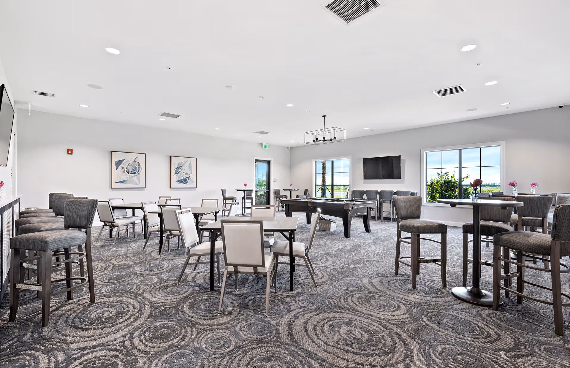 A spacious community room with multiple tables and chairs arranged for seating. The room features a patterned carpet, a pool table in the center, a wall-mounted TV, and large windows letting in natural light. There are modern light fixtures on the ceiling and abstract artwork on the walls.