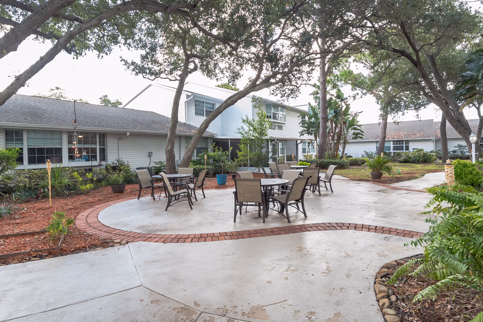 Outdoor courtyard with round paved seating area, tables and chairs surrounded by trees and low two-story buildings.
