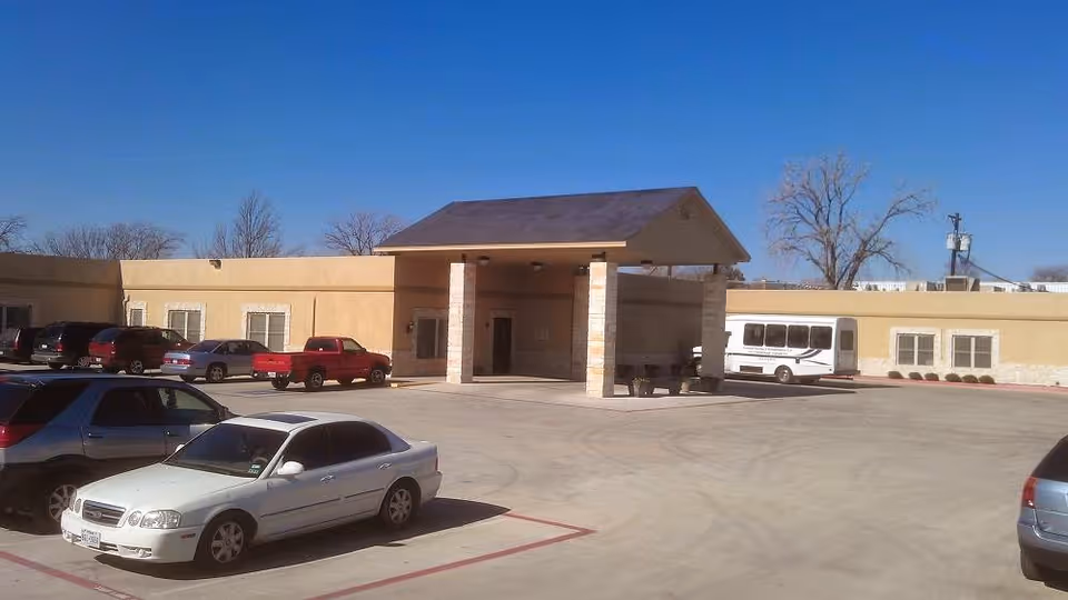 Exterior view of Garland Nursing & Rehabilitation Center showing a single-story building with a covered entrance supported by stone pillars. Several cars and a white shuttle bus are parked in the parking lot in front of the building under a clear blue sky.