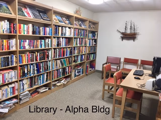 Interior view of a library room with wooden bookshelves filled with books along the left wall. There are several wooden chairs with red cushions and a wooden table with a computer and keyboard on the right side. A decorative model ship is mounted on the wall. The floor is carpeted and the room is well-lit with a ceiling light.