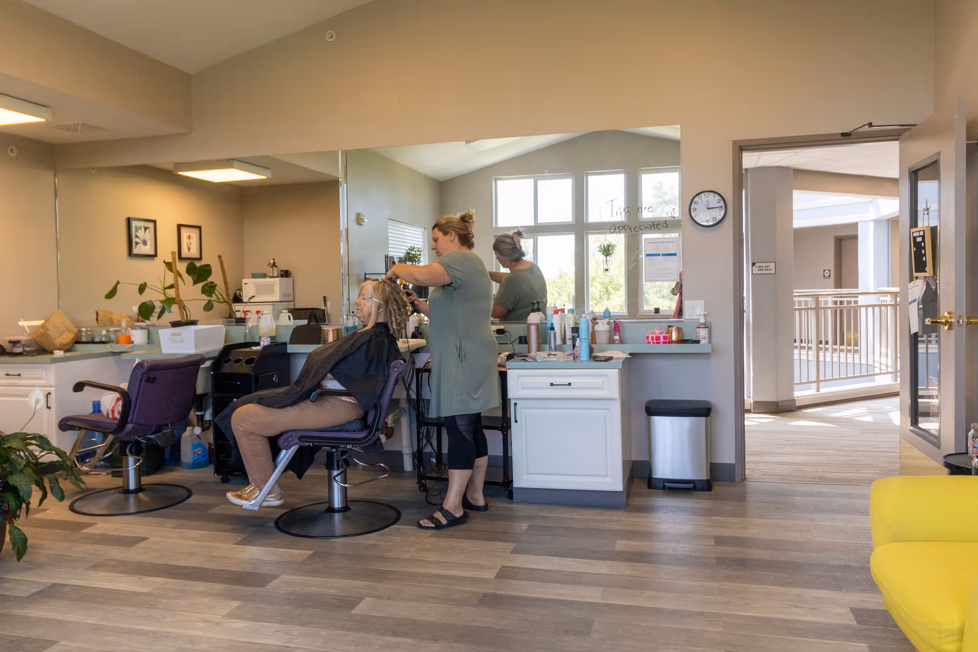 A hair salon area inside a senior living facility where a woman is seated in a purple salon chair getting her hair styled by a hairdresser. The room has large windows, a clock on the wall, various hair products on the counter, and a yellow chair in the foreground.