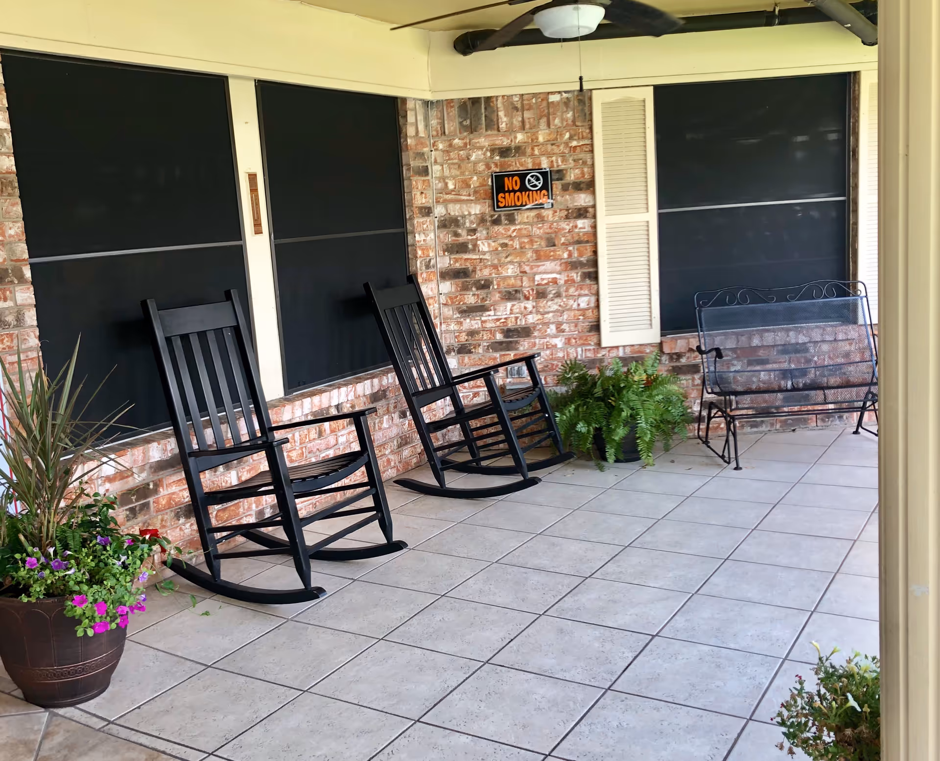 A covered outdoor patio area with two black rocking chairs, a metal bench, potted plants, and a ceiling fan. The wall is made of brick with black window coverings and a 'No Smoking' sign.