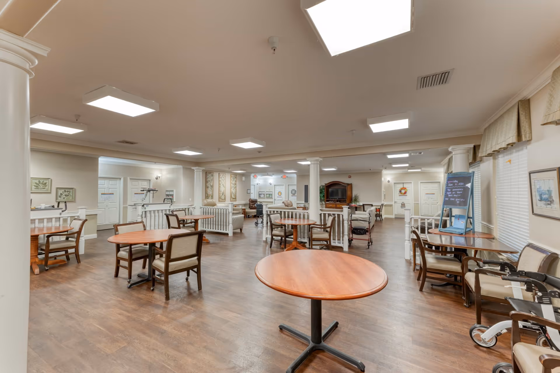 A spacious common area in a senior living facility with several round wooden tables and chairs arranged on a wood floor. The room features white columns, large ceiling lights, and windows with beige curtains. There are a few walkers and a television on a wooden stand in the background, along with some framed artwork on the walls.