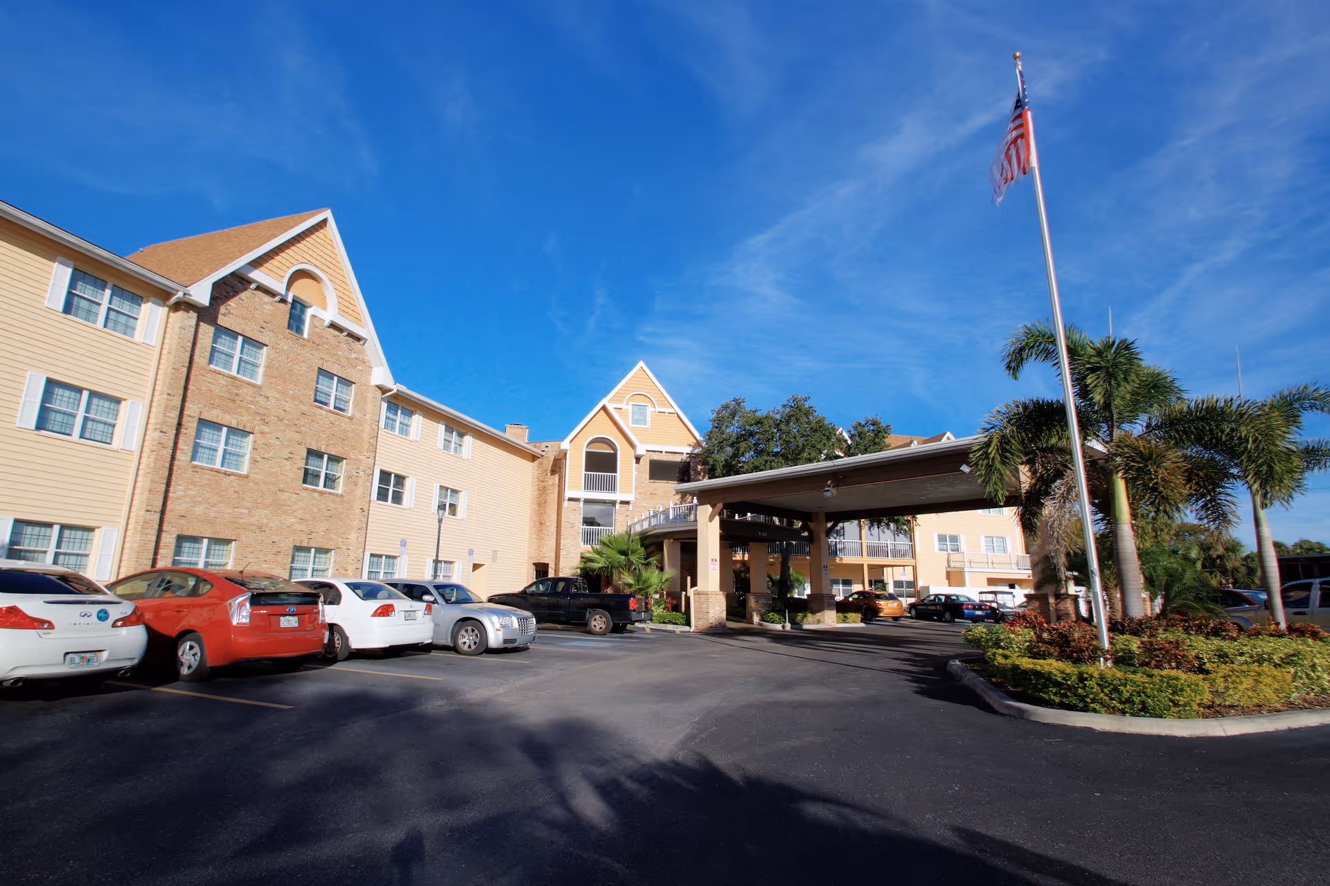 Front entrance of a multi-story senior living building with parked cars, a covered porte-cochère, palm trees, and an American flag under a blue sky.