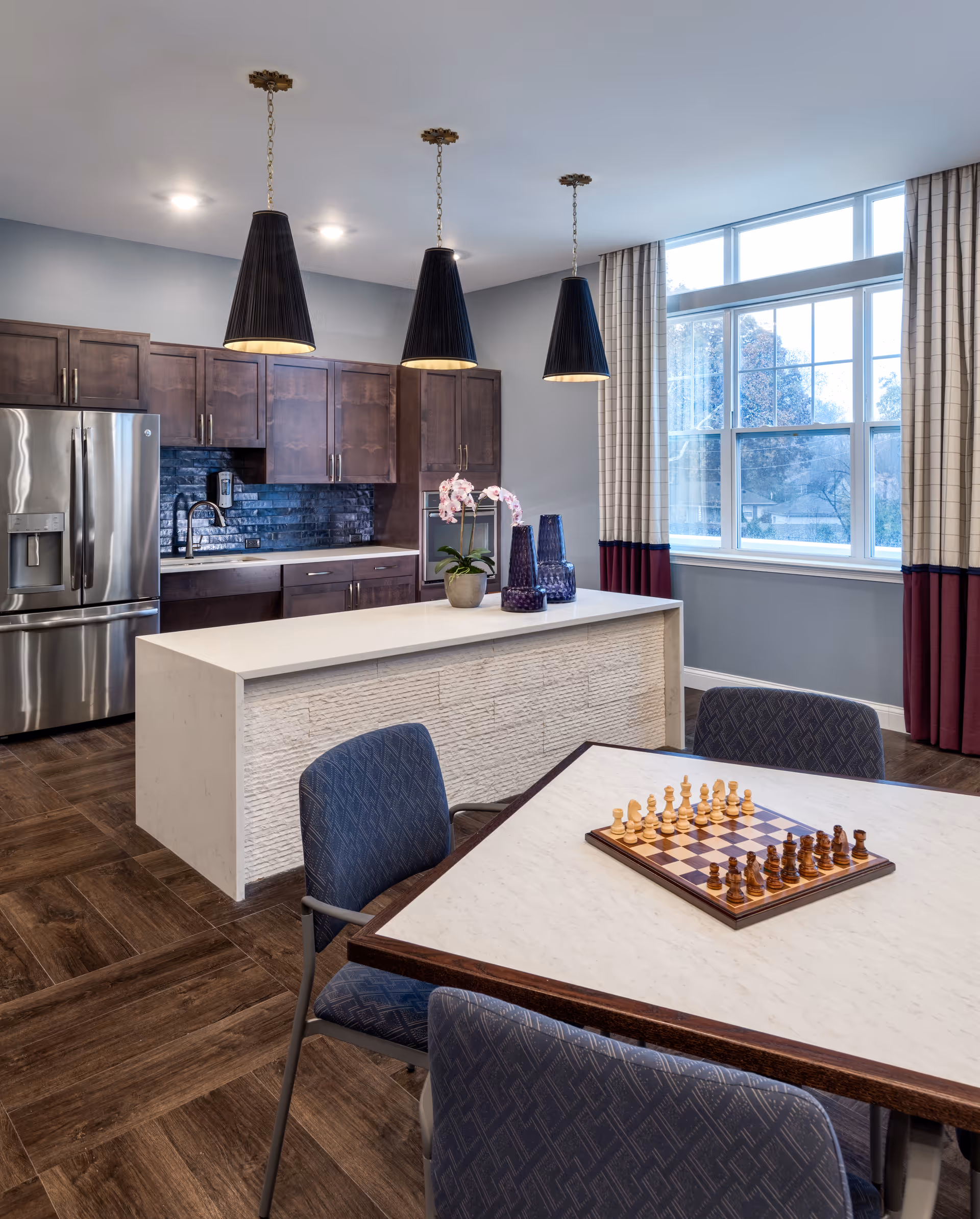 Modern kitchen area with dark wood cabinets, stainless steel refrigerator, and a white island countertop decorated with a potted orchid and two blue vases. In the foreground, a table with a chessboard set up and four blue cushioned chairs. Large window with curtains allows natural light into the room.