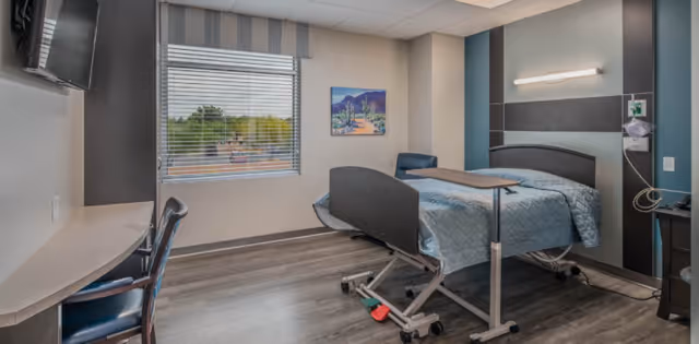 A clean and modern hospital-style bedroom with a single adjustable bed covered with a light blue quilt, a small overbed table, a chair, a wall-mounted television, and a window with blinds showing greenery outside. The room has wood-look flooring and a painting on the wall.