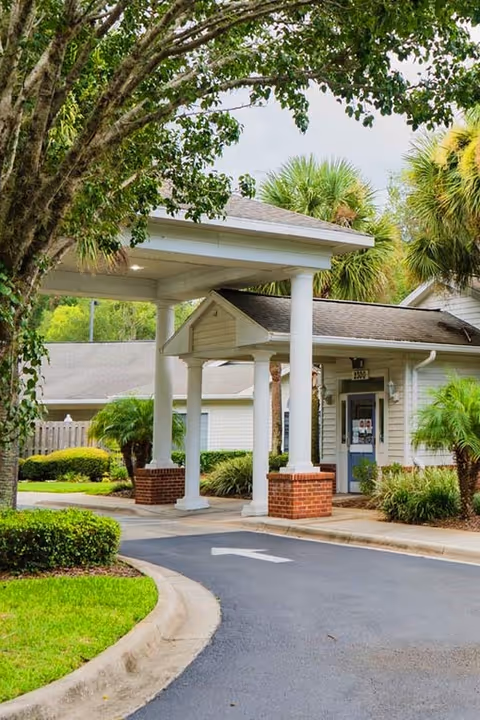 Entrance area of a senior living facility with a covered drop-off zone supported by white columns with brick bases. The building is surrounded by green trees and shrubs, with a paved driveway leading up to the entrance.