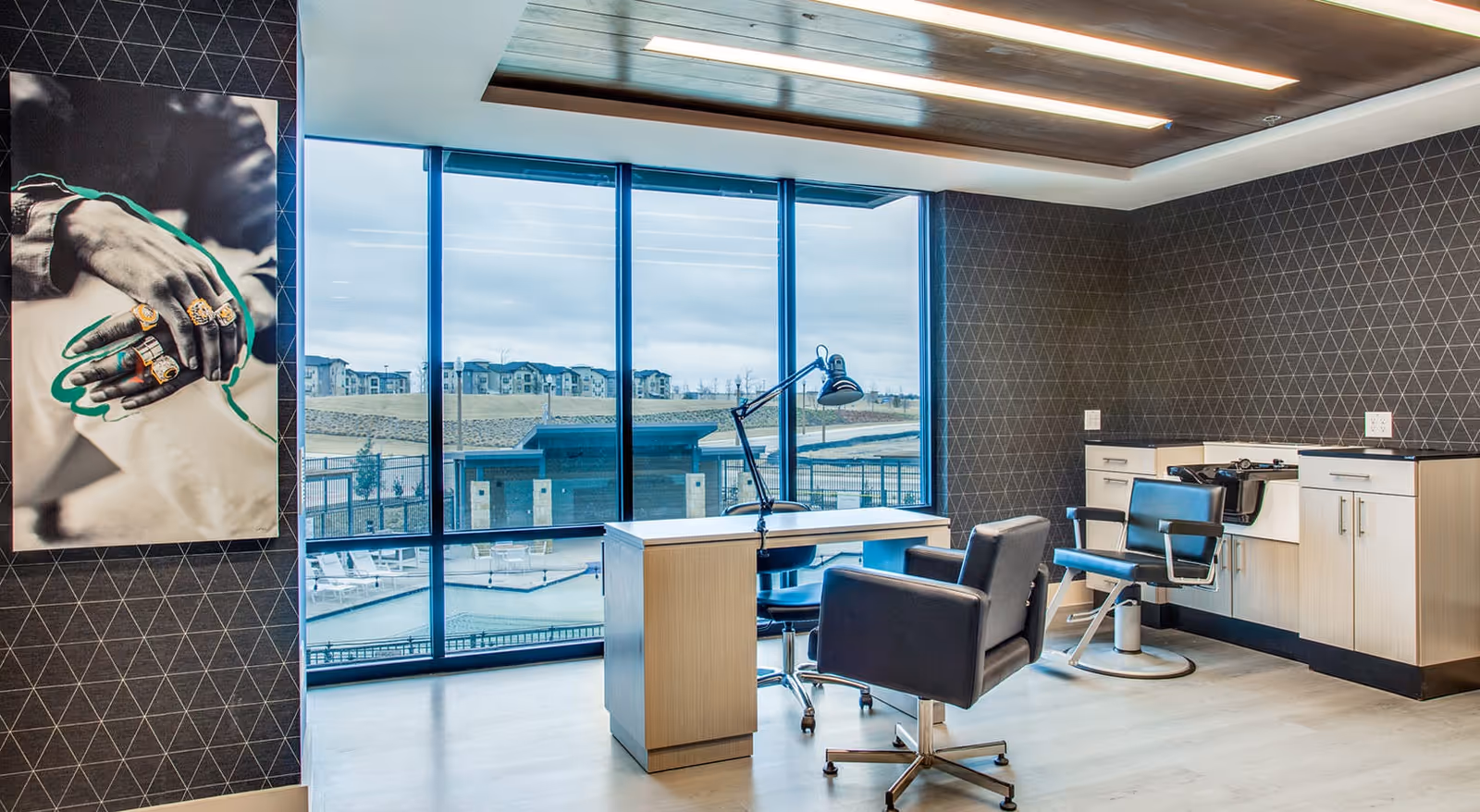 Modern salon room with two black salon chairs, a wooden desk with a desk lamp, large windows showing an outdoor view of buildings and a pool, and a black and white photo of hands with rings on the wall.