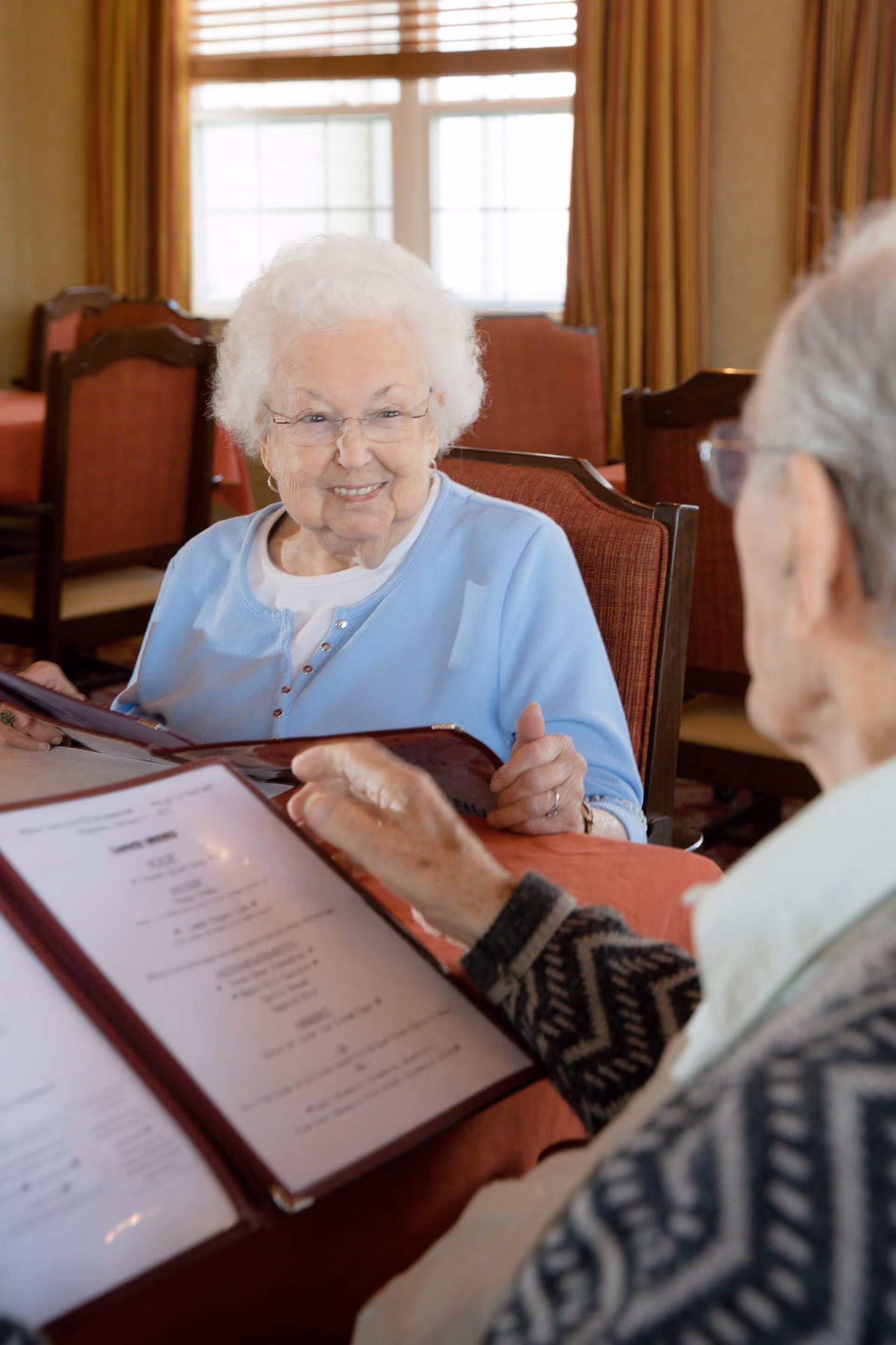 Two elderly women sitting at a table in a dining area, looking at menus. One woman with white hair and glasses is smiling, while the other woman is partially visible from behind. The room has large windows with curtains and upholstered chairs.