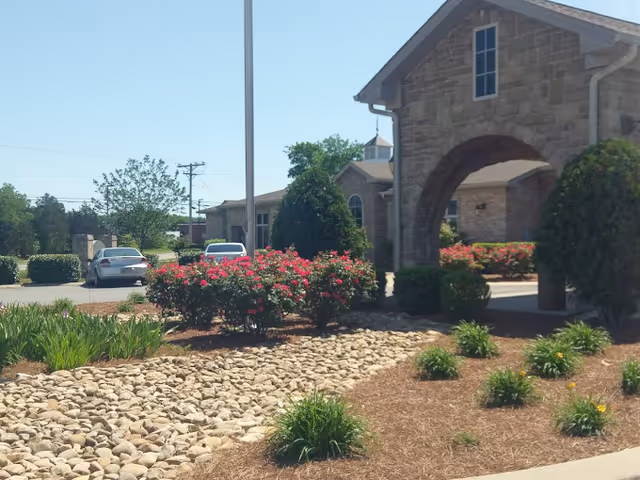 Exterior view of a senior living facility with a stone building featuring an arched entrance. In front, there are landscaped areas with bushes, flowering plants, and a rock garden. Two cars are parked in the background near the building.