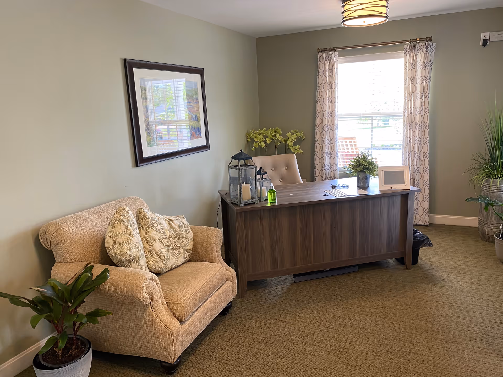 A cozy office space with a wooden desk and a cushioned chair behind it. On the desk are decorative items including a small plant, a lantern with candles, a hand sanitizer bottle, and a picture frame. To the left of the desk is a beige upholstered armchair with two patterned pillows. The room has light green walls, a window with patterned curtains, and several potted plants on the floor.