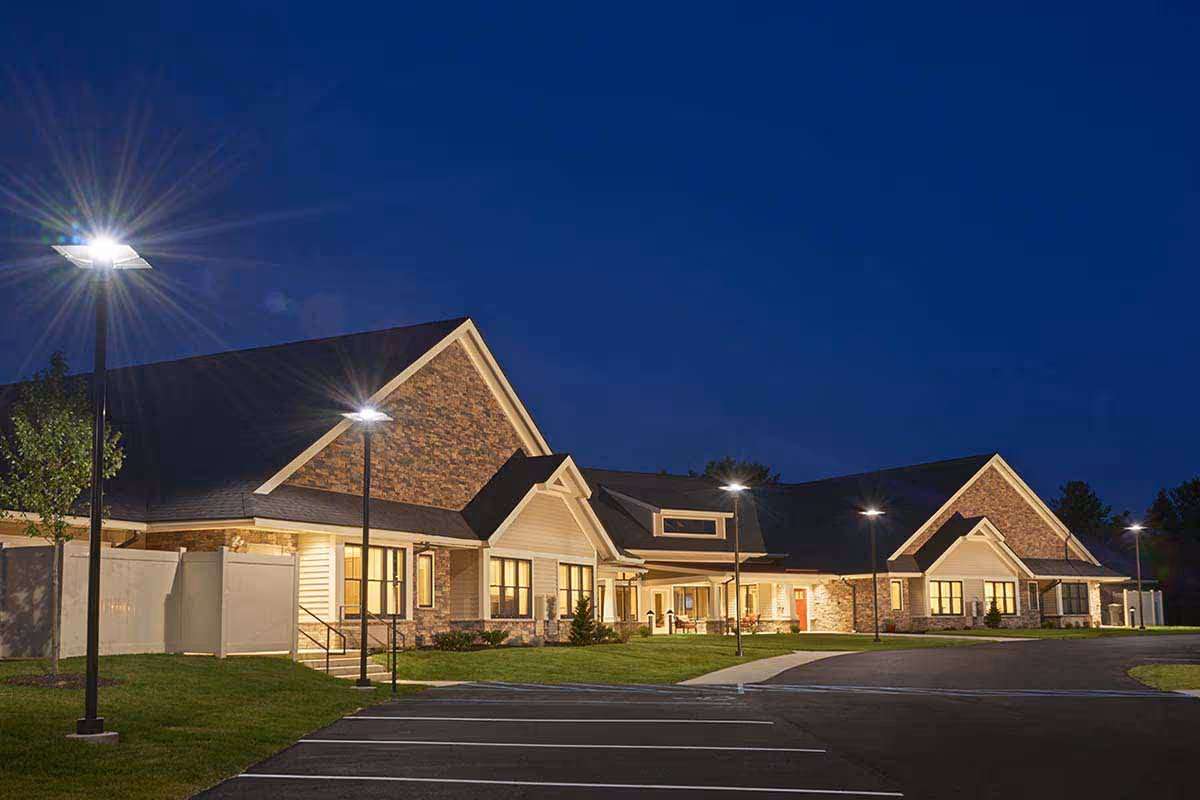 Exterior view of Legacy Place Cottages at night, showing a well-lit building with stone and siding facade, multiple windows, and a parking lot with street lamps.
