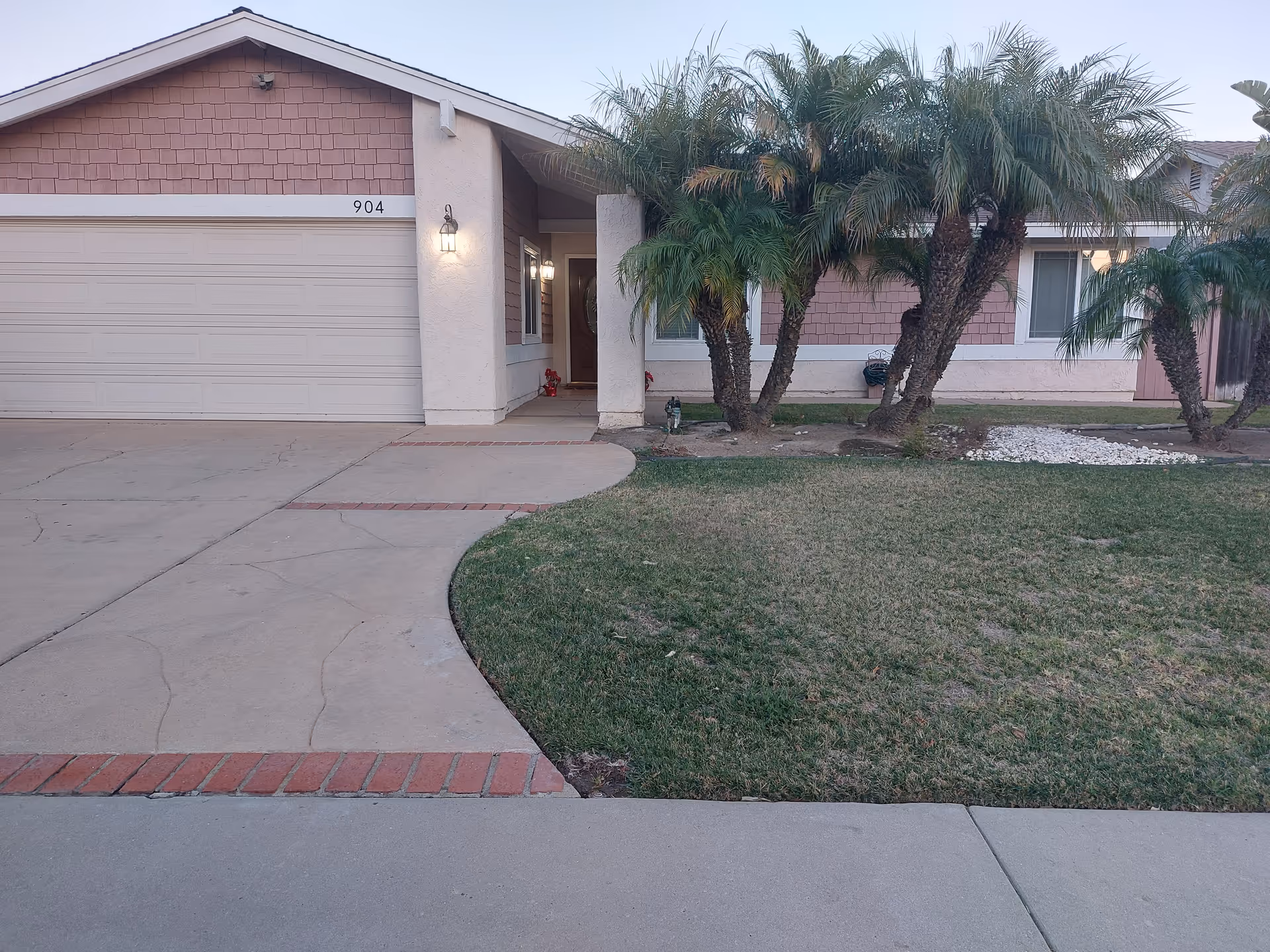 Front exterior view of a single-story residential house with a two-car garage, a concrete driveway, a small lawn with palm trees, and a covered entrance with a lit porch light.