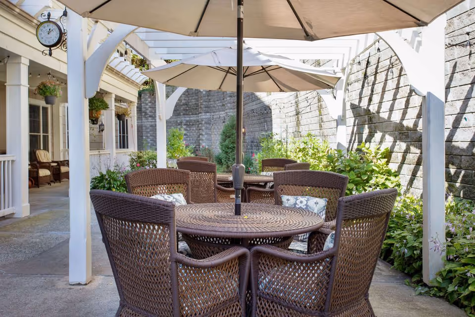 Shaded outdoor patio with round wicker tables and chairs under umbrellas and a white pergola beside a garden and building.
