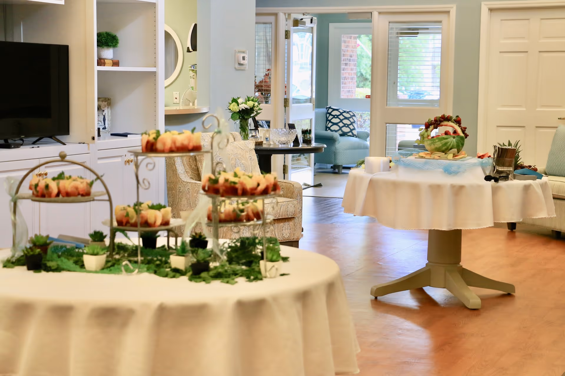 Decorated communal dining area with round tables holding tiered appetizer displays and seating in a senior living facility.