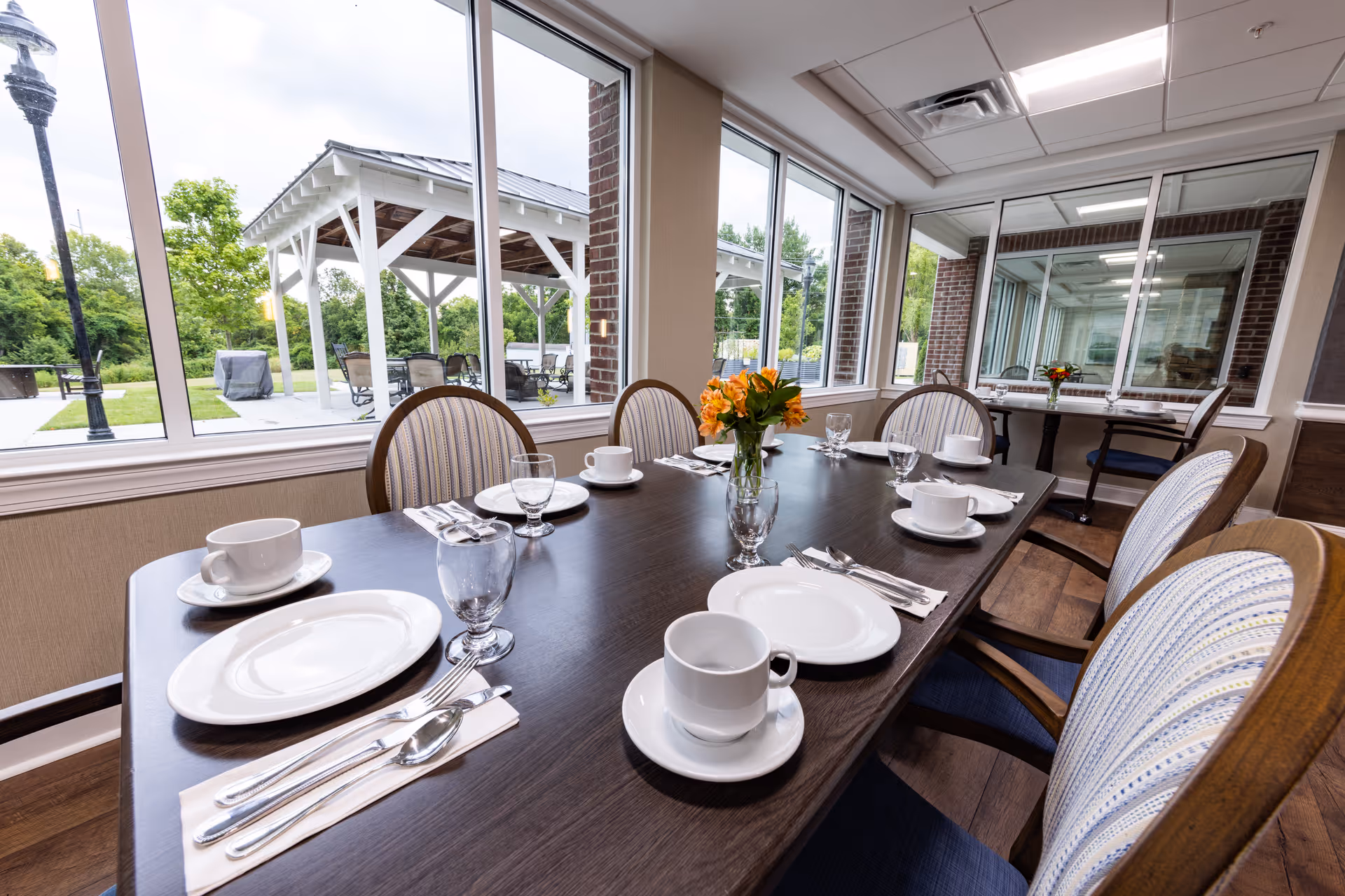 Dining room table set with plates, cups, and silverware beside large windows overlooking an outdoor pavilion.