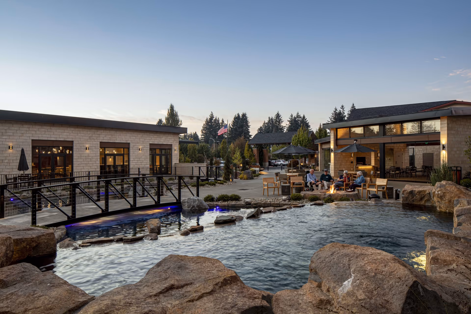 Courtyard with a pond, rock landscaping, an illuminated footbridge and people sitting by a firepit outside the facility buildings.