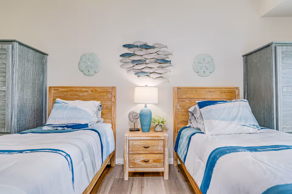 Two twin beds with blue-trim bedding flanking a wooden nightstand topped with a lamp and coastal wall decor.