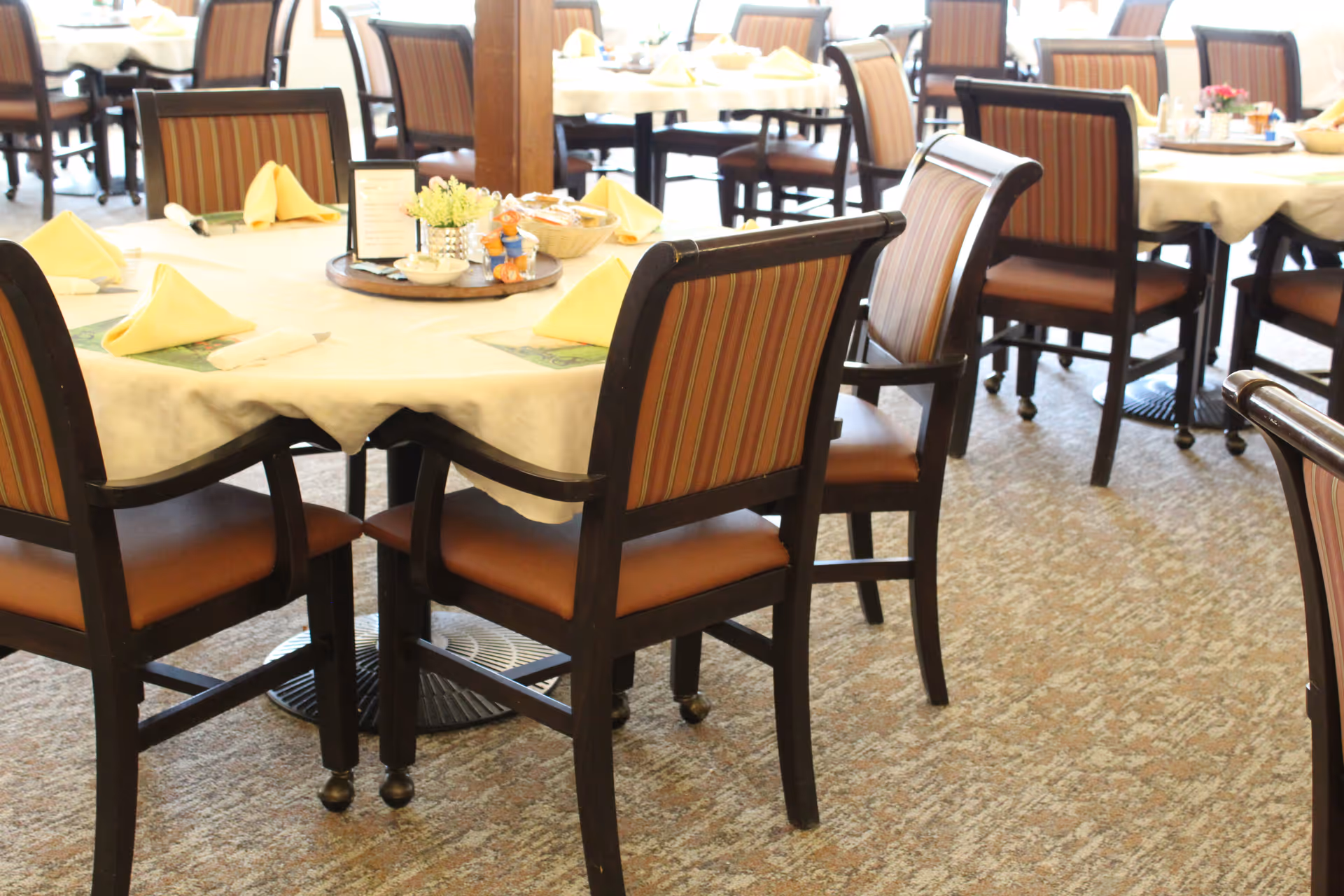 A dining area with multiple round tables covered with white tablecloths, each set with yellow folded napkins, placemats, and condiment trays. The chairs have dark wooden frames with striped upholstery on the backrests and brown cushioned seats. The floor is carpeted with a patterned design.