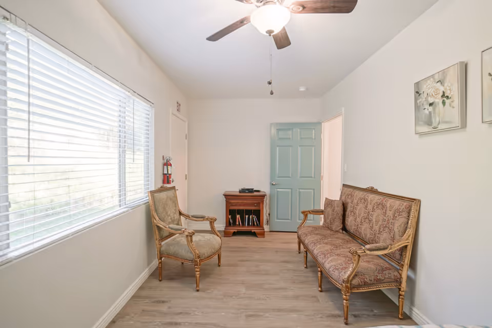 A small sitting room with a vintage upholstered sofa and matching armchair, a wooden side table with books and a device on top, a ceiling fan with light, a large window with blinds, and two floral paintings on the wall.