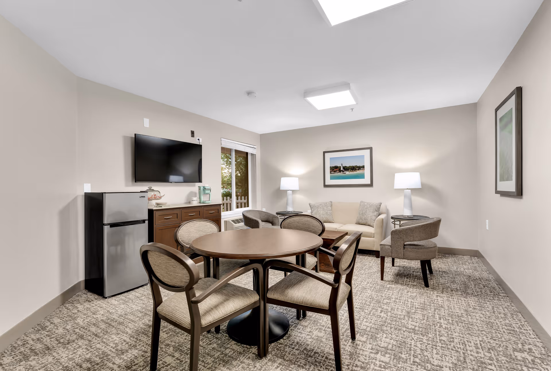 A cozy living room area in Holiday Blue Water Lodge featuring a round wooden table with four chairs, a beige sofa with two cushions, two armchairs, two side tables with lamps, a wall-mounted flat-screen TV, a small refrigerator, and a window with a view of greenery outside. The room has neutral-colored walls and carpeted flooring with framed artwork on the walls.