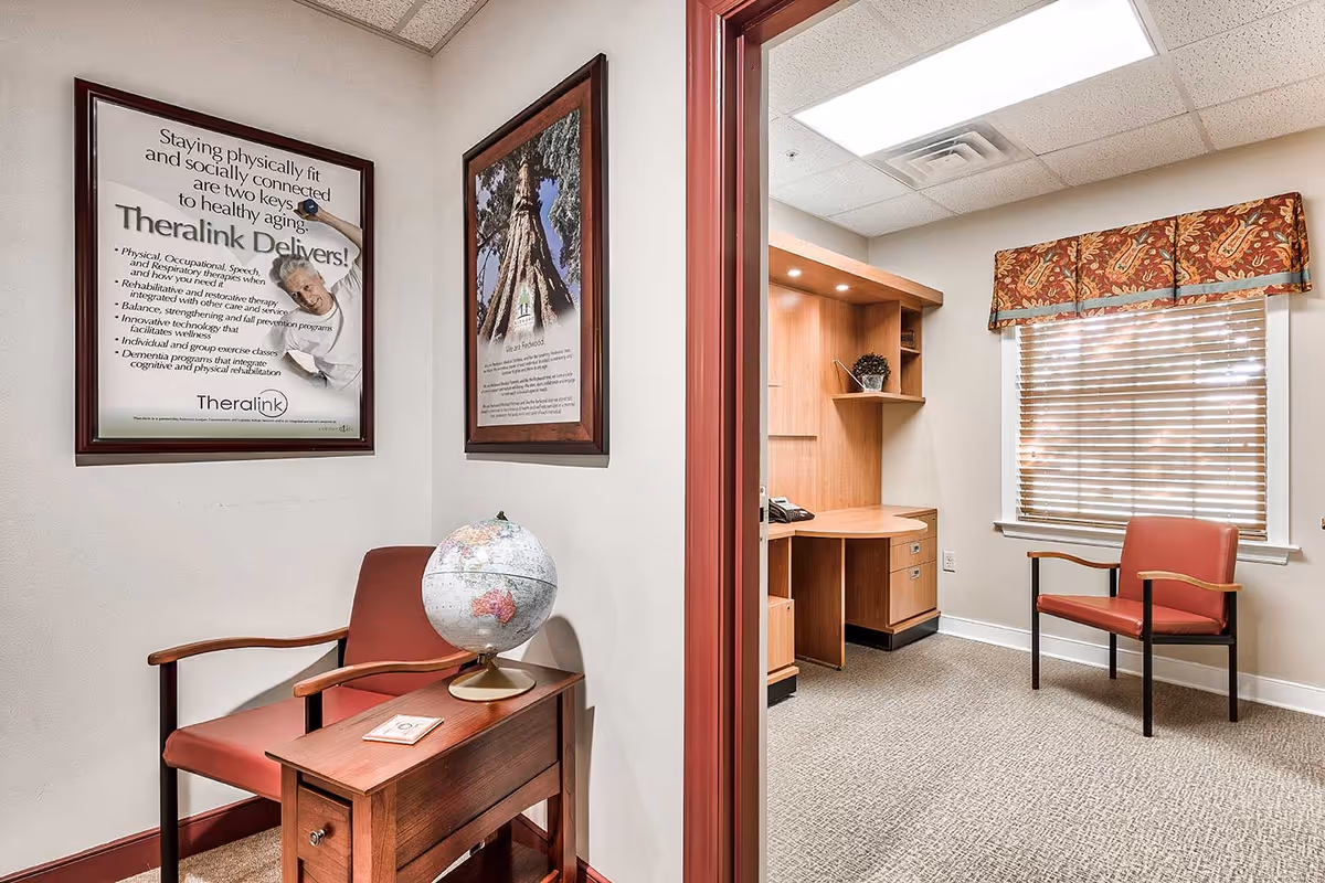 Interior view of a small office or consultation room in a senior living facility. The room features two red chairs with wooden armrests, a small wooden side table with a globe on it, framed posters on the walls, a wooden desk with built-in shelves, a telephone, and a window with blinds and a decorative valance.