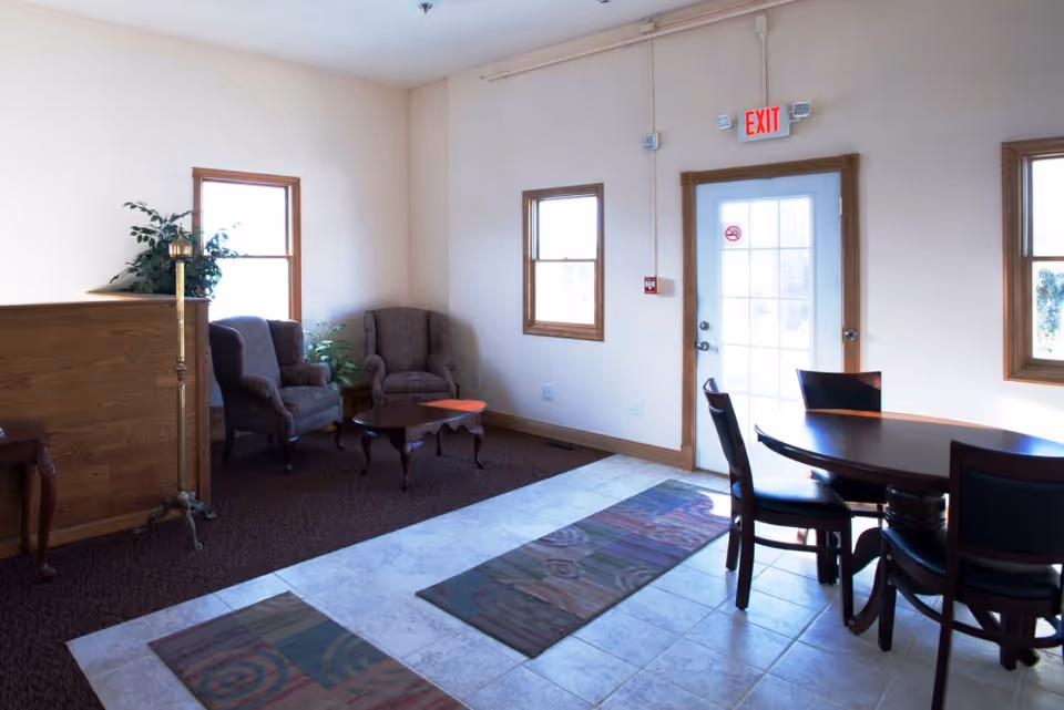 A bright room with two armchairs and a small coffee table near two windows, a round dining table with four chairs, a tiled floor with two rugs, and a door with an exit sign above it.